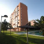 edificio residencial de varios pisos con piscina y jardín con palmeras y sombrillas de paja