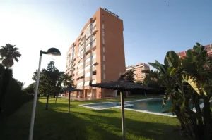 edificio residencial de varios pisos con piscina y jardín con palmeras y sombrillas de paja