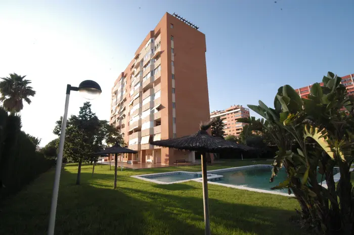 edificio residencial de varios pisos con piscina y jardín con palmeras y sombrillas de paja